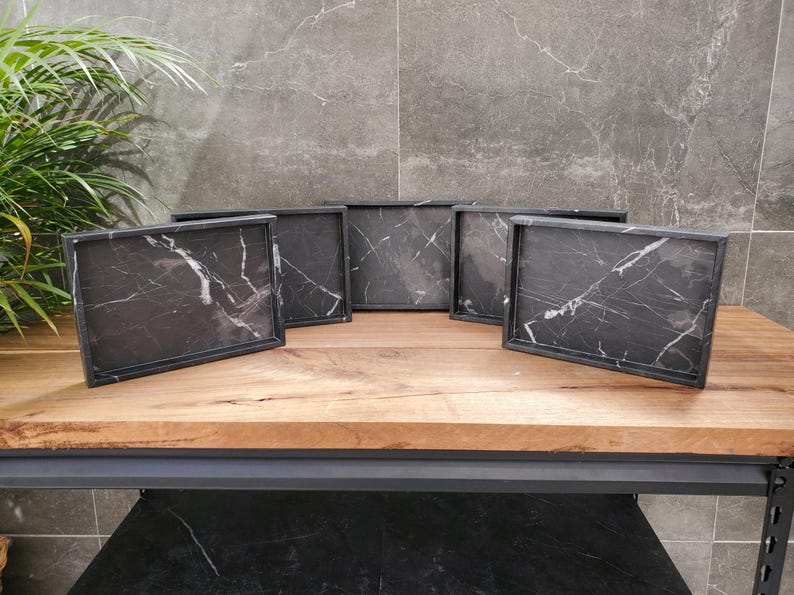 May include: Six rectangular serving trays with black marble-like surfaces and white veining are displayed on a wooden table. The trays have a dark frame and are arranged in a semi-circle. The background features a gray tiled wall and a green plant.