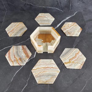 May include: A set of six hexagonal coasters and a matching holder. The coasters and holder are made of a natural stone with a layered pattern of beige, brown, and gray tones. The set is arranged on a dark gray surface.