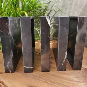 May include: A set of five black marble bookends with white veining. The bookends are rectangular with a trapezoidal shape, and are arranged in a row on a wooden surface. The background includes green plants and a gray wall.