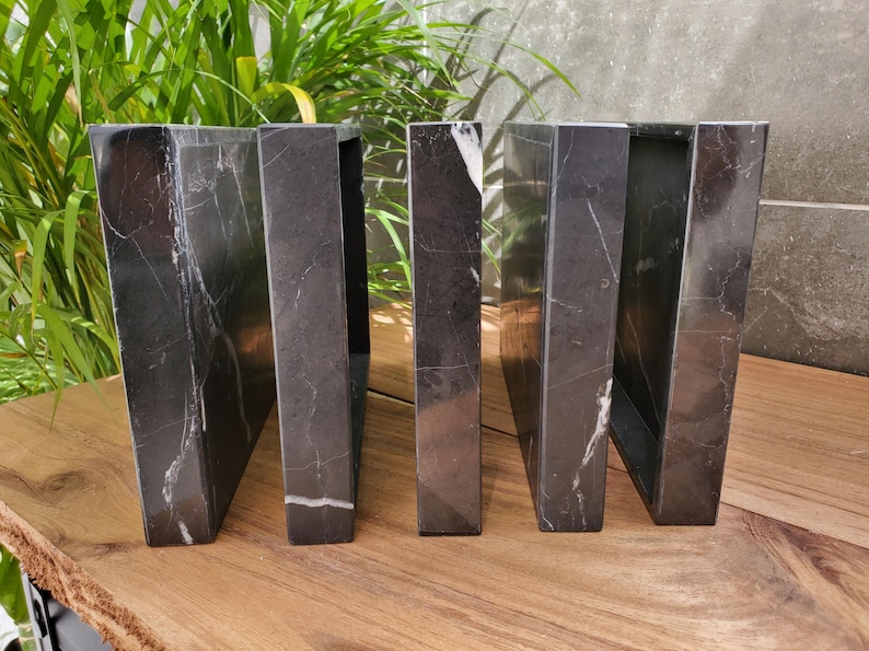 May include: A set of five black marble bookends with white veining. The bookends are rectangular with a trapezoidal shape, and are arranged in a row on a wooden surface. The background includes green plants and a gray wall.