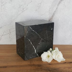 May include: A dark grey marble cube with white veining, possibly a decorative box or urn. The cube is set on a wooden surface with white flowers in the foreground. The background features a marble wall.