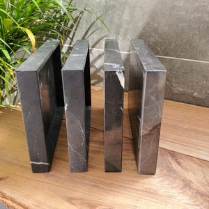 May include: Four dark gray marble bookends with white veining are arranged on a wooden surface. The bookends are rectangular with open centers, creating a modern, minimalist design. The background includes greenery and a gray wall.