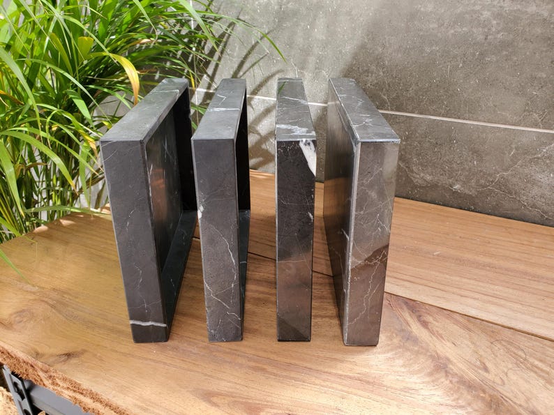 May include: Four dark gray marble bookends with white veining are arranged on a wooden surface. The bookends are rectangular with open centers, creating a modern, minimalist design. The background includes greenery and a gray wall.