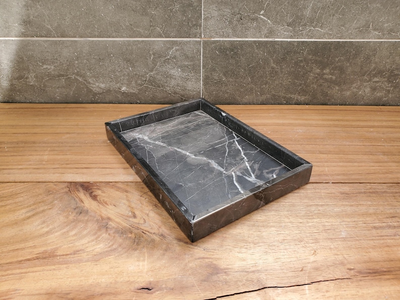 May include: A rectangular black marble tray with white veining. The tray is set on a wooden surface and has a simple, modern design. The tray is suitable for use as a decorative piece or for organizing small items.