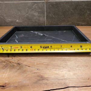May include: A rectangular black marble tray with white veining, approximately 10 inches long. The tray is on a wooden surface, with a measuring tape alongside it.