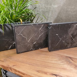 May include: Three rectangular black marble serving boards with white veining are displayed on a wooden surface. The boards vary in size and are arranged to show their different dimensions. Green plants are in the background.