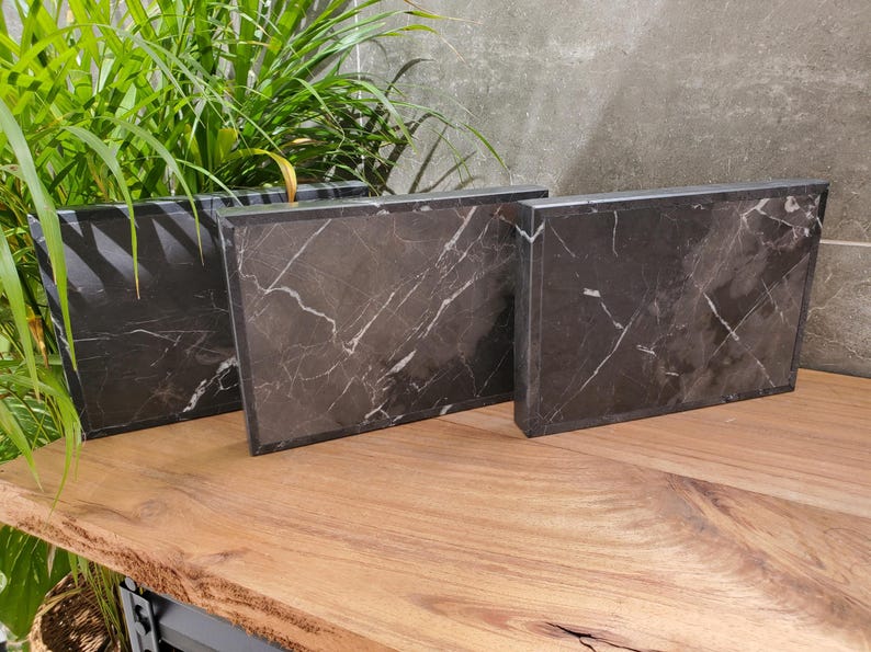 May include: Three rectangular black marble serving boards with white veining are displayed on a wooden surface. The boards vary in size and are arranged to show their different dimensions. Green plants are in the background.