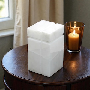 May include: A rectangular white stone container with a smooth, polished finish and a lid. The object features a vertical groove running down the center of the front and top surfaces, creating a clean, geometric appearance with subtle grey veining.