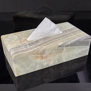 以下が含まれることがあります： 緑、グレー、ベージュの大理石で作られた長方形のティッシュボックス。白いティッシュが上部の開口部から出ています。ボックスは研磨仕上げで、暗い背景に配置されています。