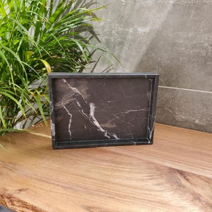 May include: A rectangular black marble tray with white veining sits on a wooden surface. The tray has a raised border and is displayed in front of a gray stone wall and green plant.