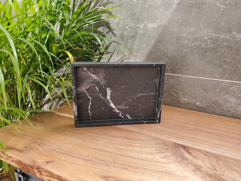 May include: A rectangular black marble tray with white veining sits on a wooden surface. The tray has a raised border and is displayed in front of a gray stone wall and green plant.