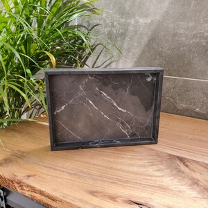 May include: A rectangular black marble tray with white veining sits on a wooden surface. The tray has a dark frame and a polished finish. The background includes a green plant and a gray wall.