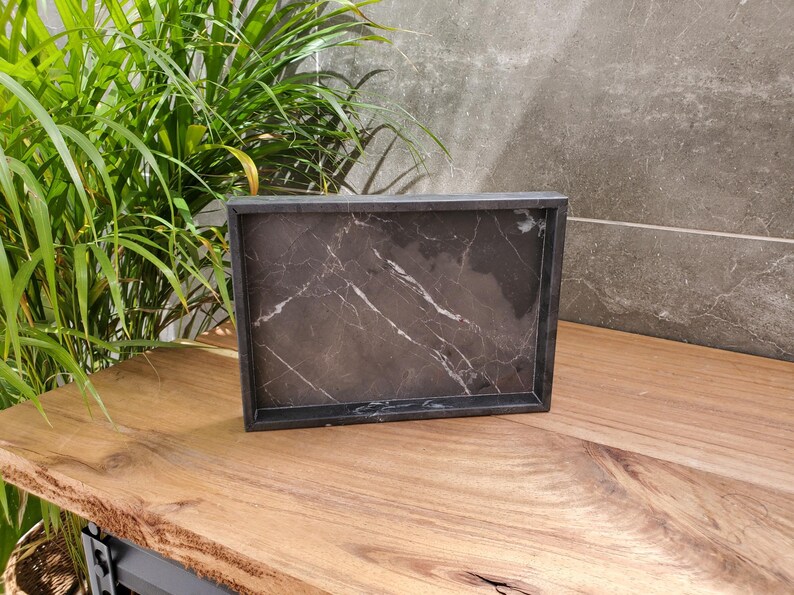 May include: A rectangular black marble tray with white veining sits on a wooden surface. The tray has a dark frame and a polished finish. The background includes a green plant and a gray wall.