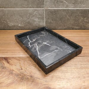 May include: A rectangular black marble tray with white veining. The tray has a raised edge and sits on a wooden surface. The background features a gray tiled wall.