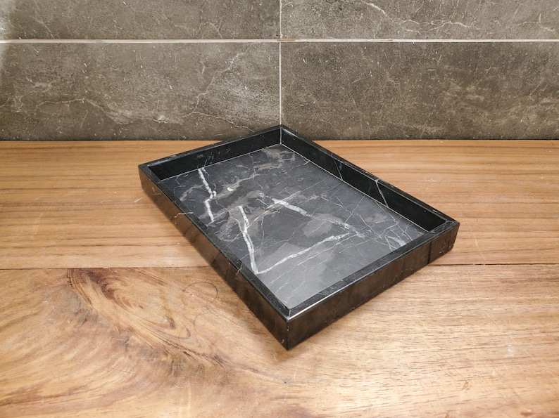 May include: A rectangular black marble tray with white veining. The tray has a raised edge and sits on a wooden surface. The background features a gray tiled wall.