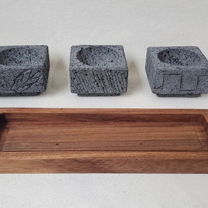 Peut inclure: Trois bougeoirs carrés en pierre grise avec des motifs sculptés, dont des feuilles et des motifs géométriques, sont placés au-dessus d'un plateau rectangulaire en bois. Le plateau est en bois brun foncé et possède une zone encastrée.