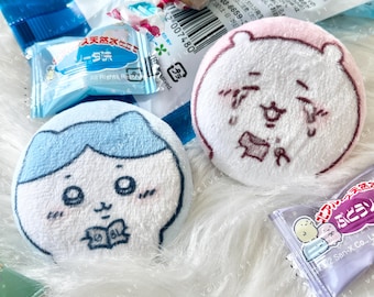 May include: Two plush, round character toys with embroidered faces, one blue and one pink, surrounded by colorful candies and packaging. The toys have detailed facial expressions and are set against a fluffy white background.