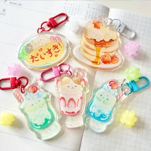 以下が含まれることがあります： キュートな食べ物をテーマにしたデザインの、透明アクリルキーホルダーのコレクション。キーホルダーには、シロップをかけたパンケーキ、パフェ、日本語のテキストが入った料理が含まれています。各キーホルダーはカラフルなデザインで、金属製の留め金が付いています。