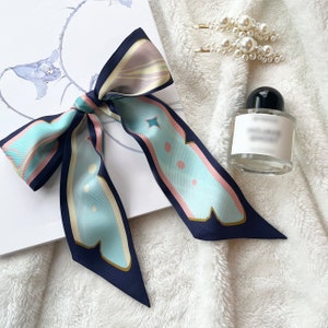 May include: A blue and green patterned silk scarf tied in a bow. The scarf has a light blue background with dark blue, pink, and cream accents. The scarf is tied around a white surface.