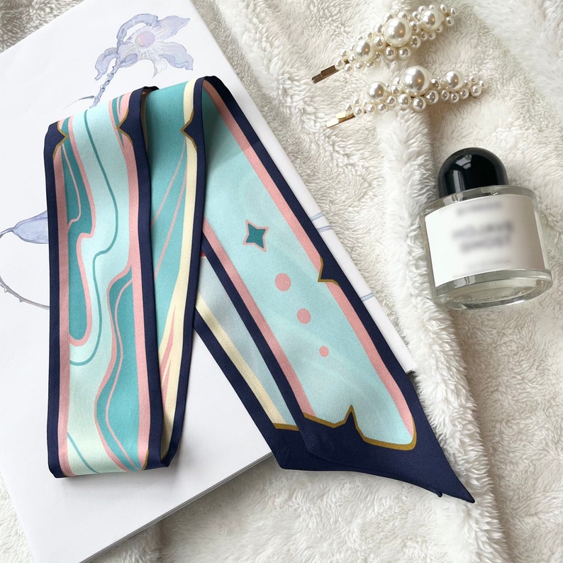 May include: A blue, pink, and white patterned silk scarf with a geometric design. The scarf is folded in half and has a navy blue trim.