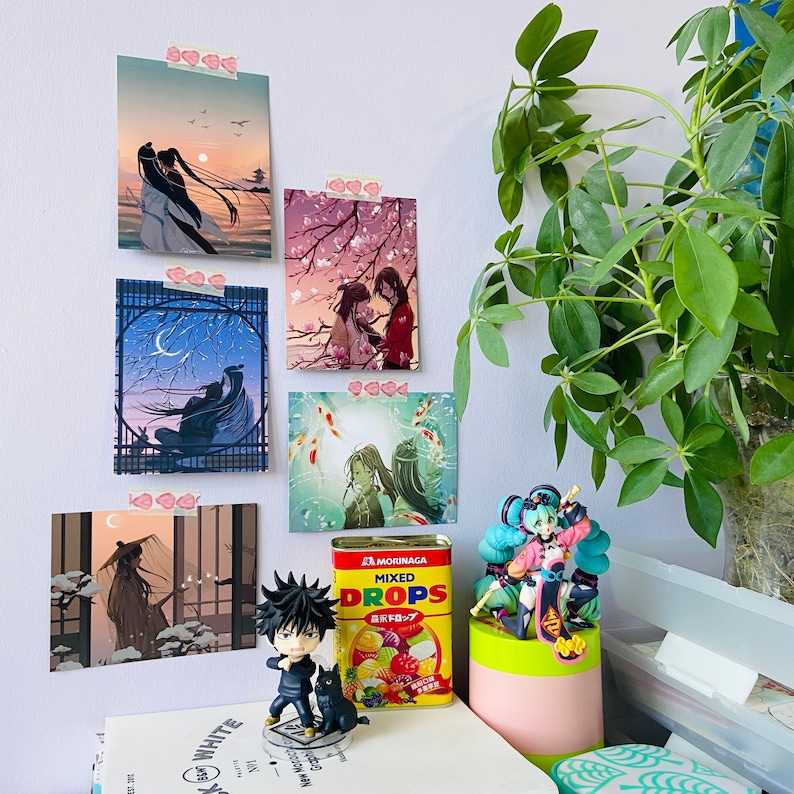 May include: A wall decorated with anime-style artwork, featuring characters in various poses and settings. The artwork is framed and hung on a white wall. A small figurine of a character with blue hair and a green dress is in the foreground. The figurine is sitting on a table with a can of mixed fruit drops.