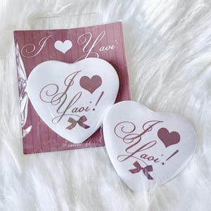 May include: Two heart-shaped white compact mirrors with the text "I 💜 Yaoi!" in a cursive font, along with a small bow and heart. One mirror is in its packaging, which has a matching design.