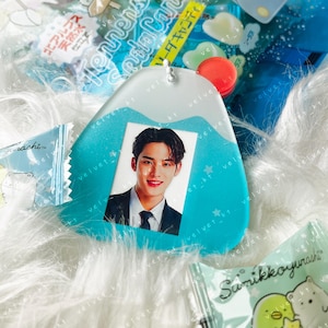 May include: A blue and white resin charm with a photo of a man with dark hair and a white shirt. The charm has a silver chain attached to it.