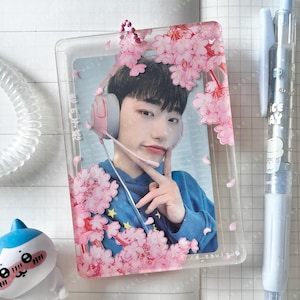 Op de afbeelding: Een doorzichtige telefoonhoes met een foto van een persoon met een headset, versierd met roze kersenbloesemmotieven. De persoon draagt een blauwe hoodie. Een pen en een klein beeldje zijn ook zichtbaar.