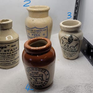 May include: Four vintage ceramic jars with blue and white labels. The jars are labeled "Bagie Jet Stain", "Cumbricks Cumberland Dairy Co. Pure", "Virol Preparation of Bone Marrow", and "Railwood's Rich Cream Honey Butter".