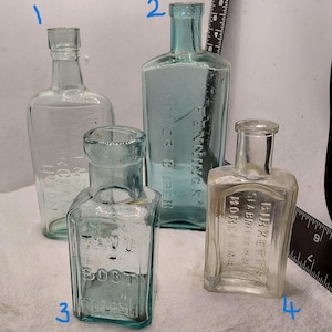 May include: Four antique glass bottles of different shapes and sizes. One bottle has the text "K & CHA IRTON" embossed on it. Another bottle has the text "PENNING'S FEVER CURE" embossed on it. The third bottle has the text "TAVU BOOT POLISH" embossed on it. The fourth bottle has the text "BIRKETTS STABRE CAMBRIDGE" embossed on it.