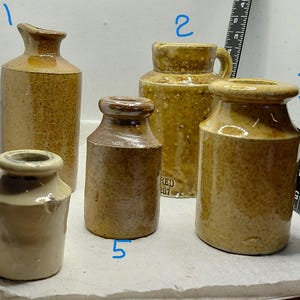 May include: Five antique stoneware jars in shades of brown and tan. The jars vary in shape and size, with textured surfaces. The jars have a rustic, vintage aesthetic. The jars are arranged on a white surface.