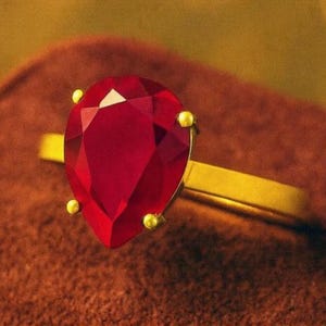 May include: A gold ring with a large, pear-cut, faceted red gemstone. The gemstone is secured by four small gold prongs. The ring band is plain, polished gold.