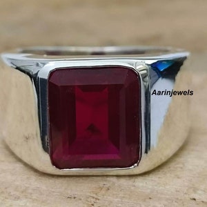 Puede incluir: Primer plano de un anillo de plata con una gran gema rectangular de color rojo intenso. El anillo tiene un acabado pulido y brillante. La gema está engastada en un bisel. El texto "Aarinjewels" es visible.
