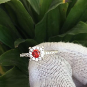 May include: A silver ring with a red gemstone surrounded by smaller clear gemstones.