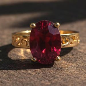 May include: A gold ring featuring a large, oval-cut, deep red gemstone. The ring's band has an openwork pattern. The gemstone is secured by four small gold prongs.