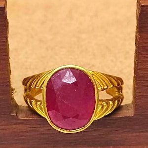 May include: A gold ring with a large, oval-shaped, faceted ruby gemstone. The ring band is textured with a braided design.