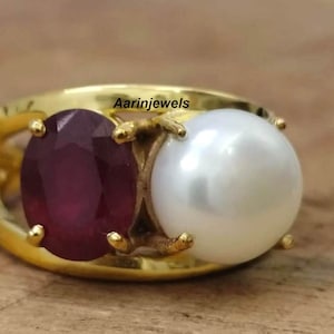 Pode incluir: Um anel de ouro com uma grande pérola branca e um rubi vermelho. O anel é adornado com uma banda de ouro e tem um design delicado.