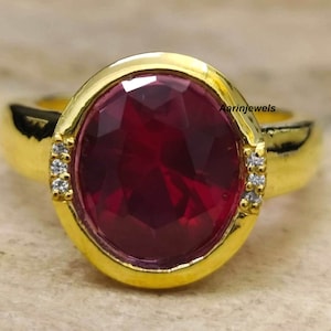 May include: A gold ring with a large, oval-shaped, red gemstone surrounded by smaller, clear gemstones. The ring is set in a gold band.
