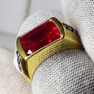 May include: A gold ring with a large, rectangular, deep red gemstone. The ring has a textured band and small blue gemstones on the sides. A bold piece of jewellery with a striking contrast.