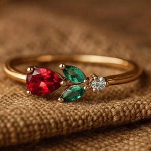 May include: A gold ring featuring a pear-shaped red gemstone, two green leaf-shaped gems, and a small white diamond. The ring is set on a textured brown surface, showcasing the intricate design and vibrant colors of the jewelry.
