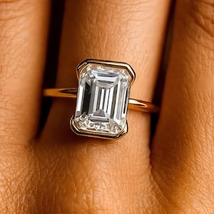 Op de afbeelding: Een gouden ring met een grote, rechthoekige, heldere edelsteen in een gouden bezelzetting. De ringband is dun en goudkleurig. De edelsteen heeft een trapsgewijze slijpvorm, die het licht reflecteert. De ring wordt aan een vinger getoond.