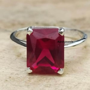 May include: A silver ring featuring a large, rectangular, deep red gemstone. The gemstone is set with four prongs, and the band is smooth and polished. The ring is displayed on a light-colored surface. The text "Aarinjewels" is visible.