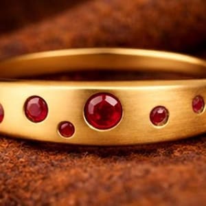 Op de afbeelding: Een goudkleurige ring met een reeks ingezette rode edelstenen. De ring heeft een platte band met gelijkmatig verdeelde cirkelvormige zettingen die de rode stenen vasthouden. De ring wordt gepresenteerd op een getextureerd bruin oppervlak.