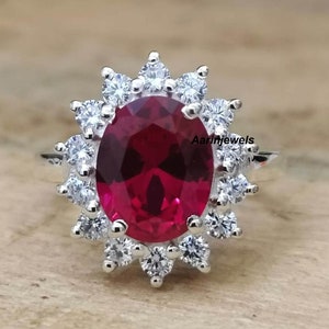 Könnte beinhalten: Ein silberner Ring mit einem großen ovalen roten Edelstein, umgeben von kleineren weißen Edelsteinen. Der Ring ist im Halo-Stil gefasst.