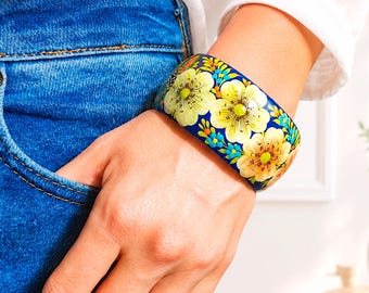 Könnte beinhalten: Ein breiter Armreif mit einer leuchtend blauen Grundfarbe, verziert mit einem sich wiederholenden Muster aus großen gelben Blumen, kleinen hellblauen Blüten und grünen Blättern, getragen am Handgelenk zu blauem Jeansstoff und einem weißen Hemd.