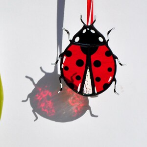 May include: A stained glass ladybug ornament featuring a vibrant red body with black spots and a black head. The piece is suspended by a thin red ribbon and displays a white textured section near the base, with black metal framing the glass.
