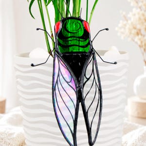Könnte beinhalten: Ein Zikaden-Ornament aus Buntglas mit einem leuchtend grünen Thorax, hellroten Augen und irisierenden, durchscheinenden Flügeln mit schwarzen Bleirändern. Die Insektenfigur hat dünne schwarze Drahtbeine, die vom Körper abstehen.