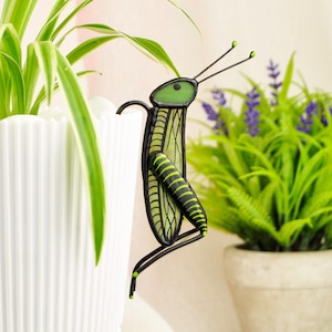 May include: A decorative metal grasshopper plant stake. The grasshopper has a green head and body with black and yellow stripes. It is perched on the edge of a white potted plant with green and white striped leaves. A second plant with purple flowers is in the background.