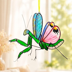 May include: A colorful, stained-glass-style praying mantis ornament. The insect features a green body, pink and blue wings, and a yellow hanging ribbon. The wings have intricate details and patterns, adding to the decorative appeal. This unique piece is perfect for nature lovers.