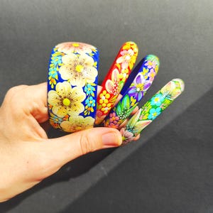 May include: A collection of hand-painted wooden bangles in various colors, featuring floral designs. The wide blue bangle displays large yellow flowers, while the others showcase smaller blooms in red, purple, and green. The bangles are held against a dark background.